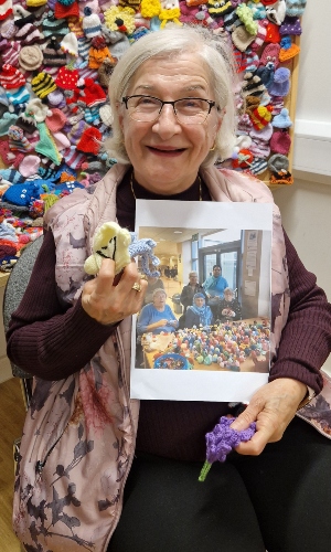 An older lady with glasses and grey hair holds up some miniature beanie hats and a printed out photo of her friend